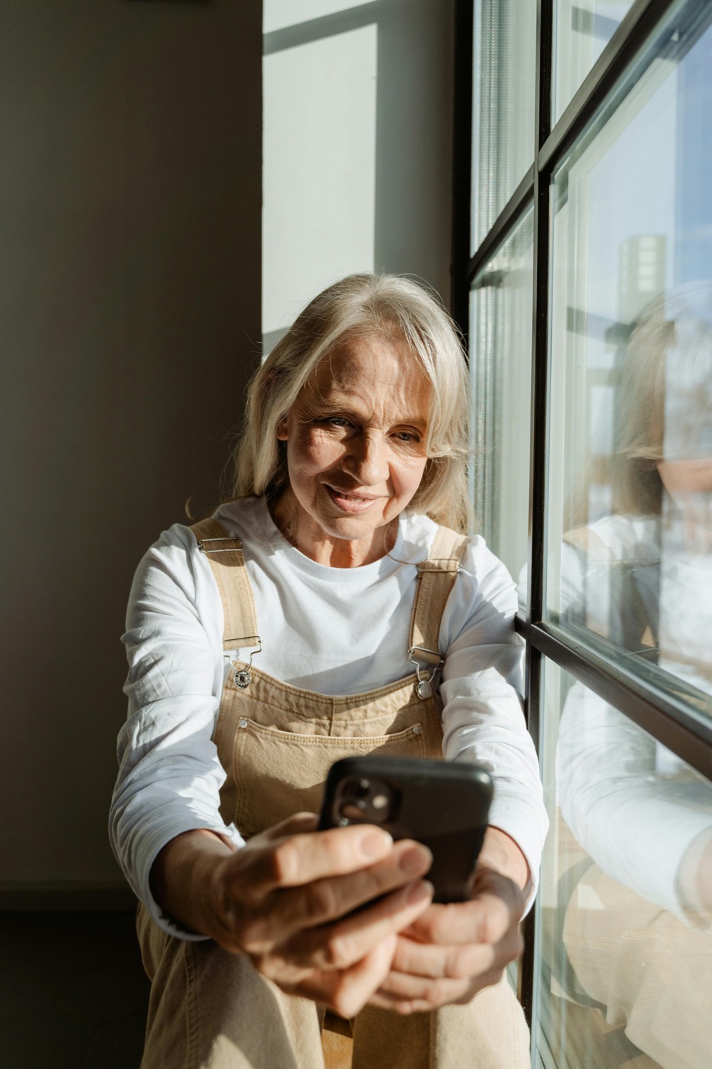 An older woman smiling while using her smartphone beside a sunny window